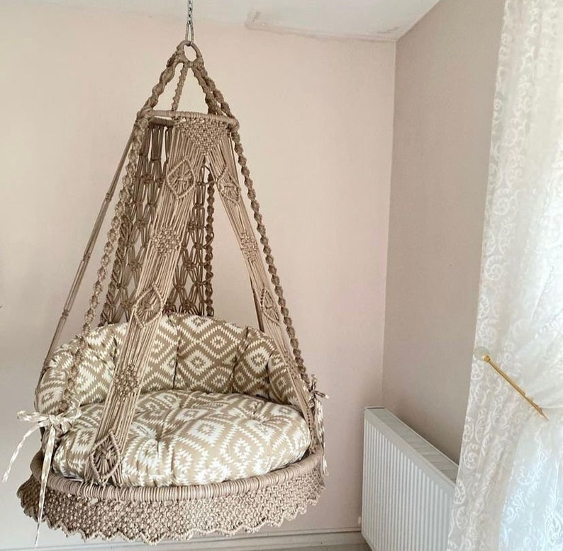 Pu&ograve; includere: Una sedia a dondolo in macram&egrave; beige con un cuscino a motivi bianchi e beige. La sedia &egrave; sospesa al soffitto con una catena metallica.