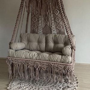 Peut inclure: Un fauteuil suspendu en macramé marron avec une bordure en franges et un coussin épais. Le fauteuil est suspendu à un crochet de plafond et est suspendu au-dessus d'un tapis marron.