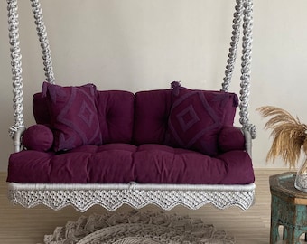 Könnte beinhalten: Eine violette gepolsterte Schaukel mit einem weißen Seil und einem gehäkelten Teppich darunter. Die Schaukel hat zwei Kissen mit geometrischen Mustern.