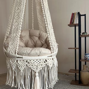Op de afbeelding: Een crèmekleurige macramé hangstoel met een gecapitonneerd beige kussen. De stoel heeft een gefranjerde onderkant en hangt aan het plafond. De kamer heeft een neutraal kleurenpalet.