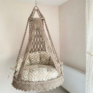 Pu&ograve; includere: Una sedia a dondolo in macram&egrave; beige con un cuscino a motivi bianchi e beige. La sedia &egrave; sospesa al soffitto con una corda.