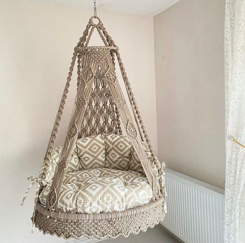 Pu&ograve; includere: Una sedia a dondolo in macram&egrave; beige con un cuscino a motivi bianchi e beige. La sedia &egrave; sospesa al soffitto con una corda.