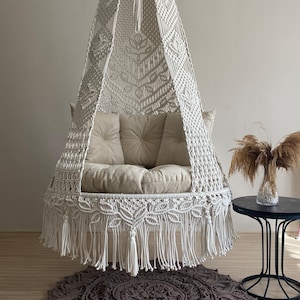 May include: A white macrame hanging chair with a fringe border and a beige cushion. The chair is hanging from the ceiling and is over a brown crocheted rug.