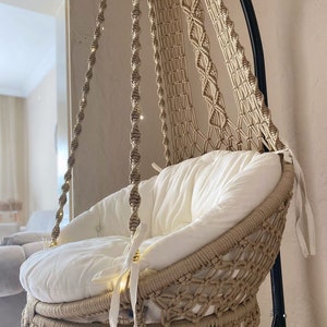 May include: A beige macrame hanging chair with a white cushion and fringe detail. The chair is suspended from a black metal stand.