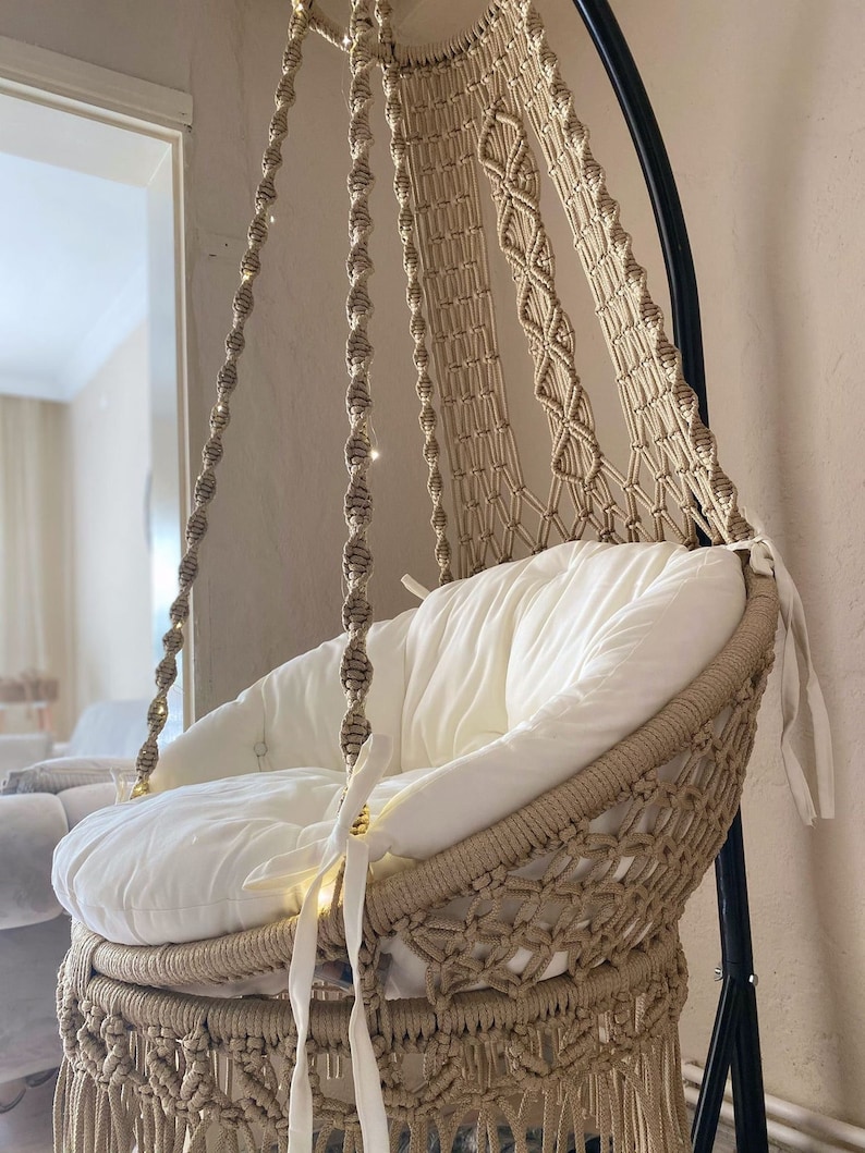 May include: A beige macrame hanging chair with a white cushion and fringe detail. The chair is suspended from a black metal stand.