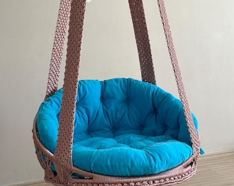 Könnte beinhalten: Ein Hängesessel mit einem blauen Kissen und einem rosafarbenen Seilrahmen. Der Sessel hängt von der Decke und befindet sich über einem weißen gehäkelten Teppich.