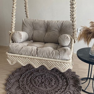 May include: A beige macrame swing chair with a gray velvet cushion and two pillows. The swing chair is hanging from the ceiling and is in front of a gray wall. A gray crocheted rug is on the floor in front of the swing chair.