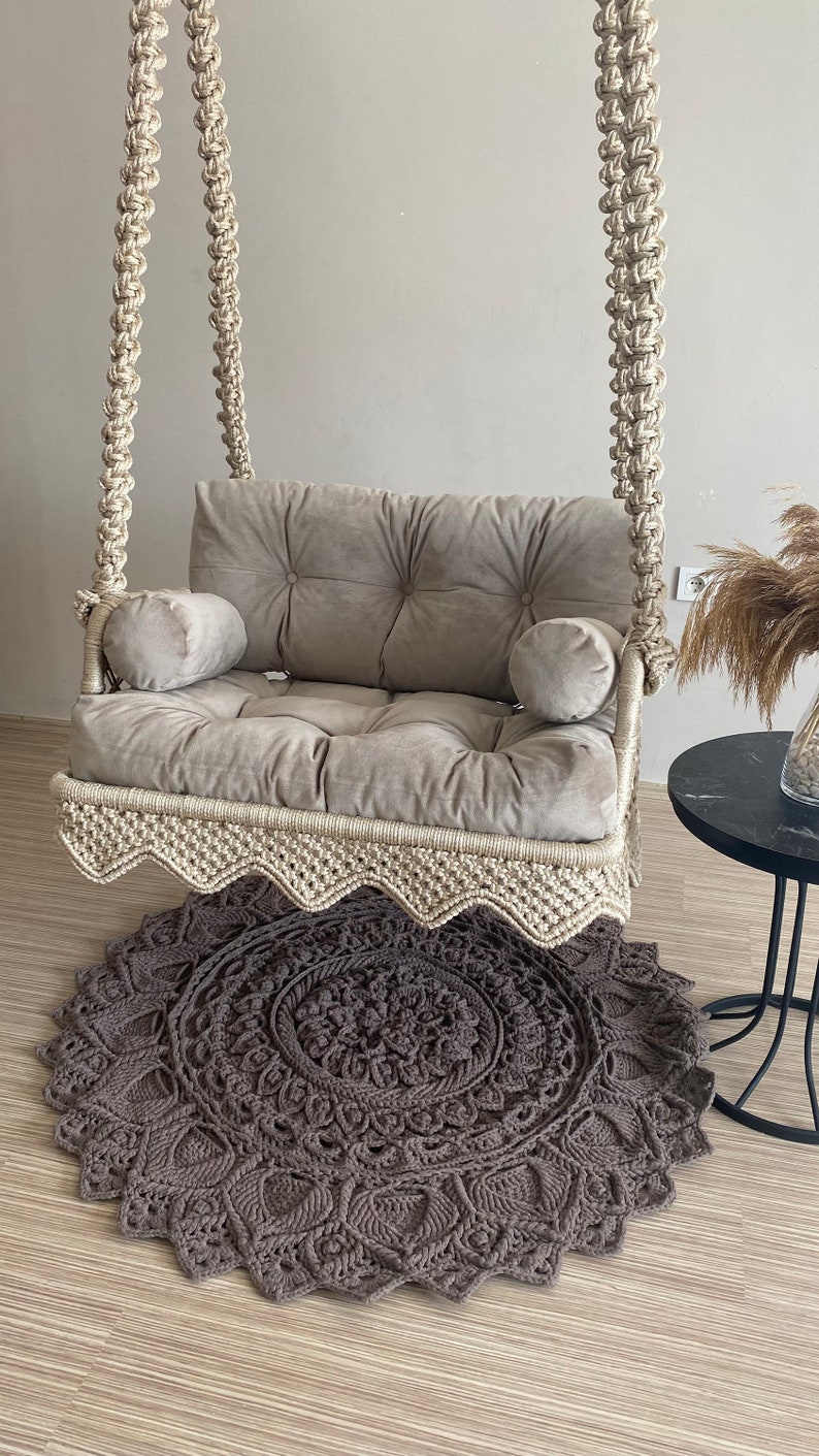 May include: A beige macrame swing chair with a gray velvet cushion and two pillows. The swing chair is hanging from the ceiling and is in front of a gray wall. A gray crocheted rug is on the floor in front of the swing chair.