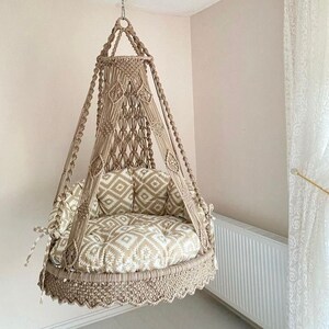 Pu&ograve; includere: Una sedia a dondolo in macram&egrave; beige con un cuscino a motivi bianchi e beige. La sedia &egrave; sospesa dal soffitto e ha un bordo a frangia.