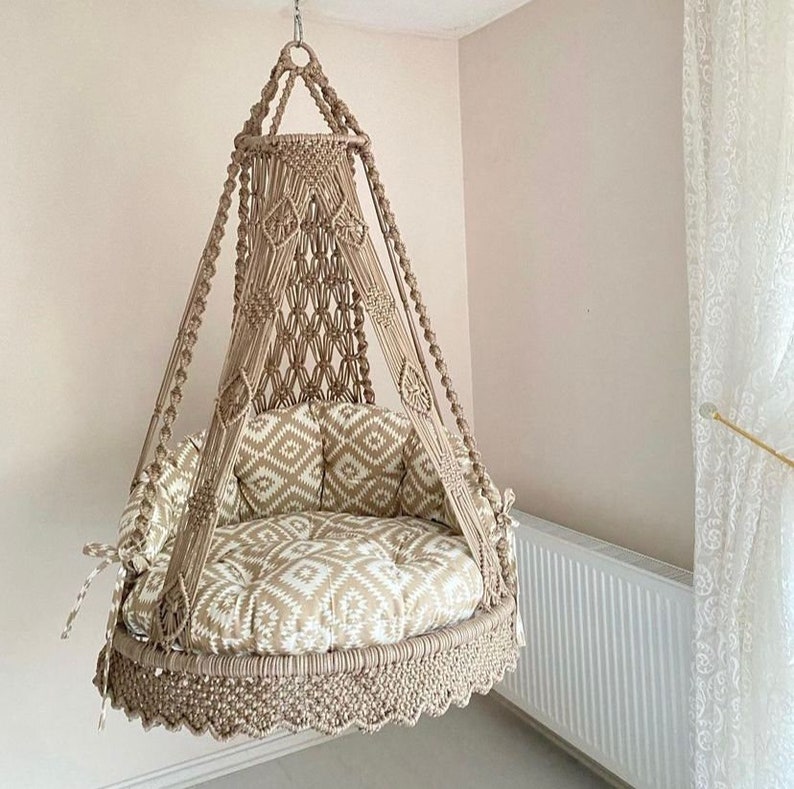 Pu&ograve; includere: Una sedia a dondolo in macram&egrave; beige con un cuscino a motivi bianchi e beige. La sedia &egrave; sospesa dal soffitto e ha un bordo a frangia.