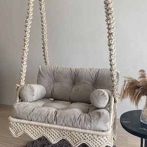 May include: A beige macrame swing chair with a gray cushion and two pillows. The swing chair is hanging from the ceiling and is over a gray and brown crocheted rug.