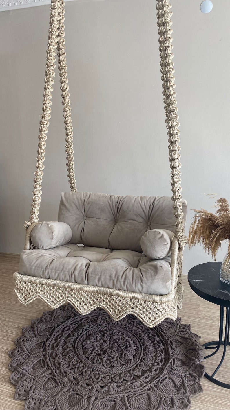 May include: A beige macrame swing chair with a gray cushion and two pillows. The swing chair is hanging from the ceiling and is over a gray and brown crocheted rug.