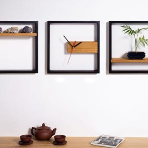 Op de afbeelding: Drie zwarte ingelijste wandplanken met houten planken. De linker plank heeft twee paarse en grijze kristallen. De middelste plank heeft een klok met zwarte wijzers en een houten gezicht. De rechter plank heeft een kleine plant in een pot.