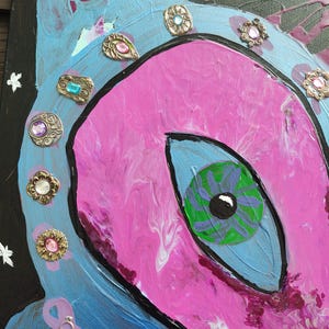 Könnte beinhalten: Ein abstraktes Gemälde in Rosa- und Blautönen mit einem großen Auge in der Mitte. Das Auge ist grün und lila mit einer schwarzen Pupille. Das Gemälde ist mit goldenen und silbernen Verzierungen verziert.