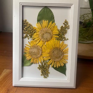 May include: A framed floral arrangement featuring three yellow sunflowers, green leaves, and small yellow flowers. The flowers are pressed and arranged on a white background within a white frame. The frame is rectangular.