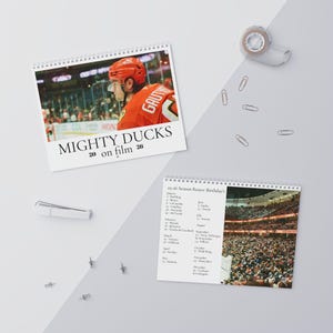 May include: Two spiral-bound calendars on a white surface. One calendar features a hockey player in an orange jersey with the text "MIGHTY DUCKS 20 on film 26." The other calendar shows a stadium crowd and a list of dates.