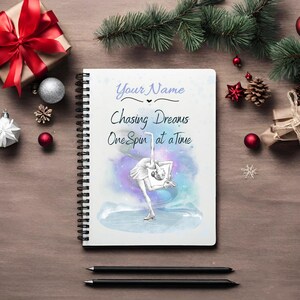 Könnte beinhalten: Ein weißes Notizbuch mit Spiralbindung und einer Aquarell-Illustration einer Eiskunstläuferin. Oben auf dem Notizbuch steht der Text "Your Name", gefolgt von "Chasing Dreams One Spin at a Time".