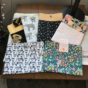 May include: Six different fabric pouches with various designs. The pouches are made of cotton fabric and have a zipper closure. The designs include a black and white pattern with shields and swords, a floral pattern, a black and white elephant pattern, a black and white pattern with small white dots, a pink and white polka dot pattern, and a pink and white floral pattern.