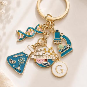 May include: Gold-coloured keyring with science-themed charms. Charms include a DNA strand, microscope, flask, and a charm with the word "CHEMISTRY". The keyring also has a charm with the letter "G".