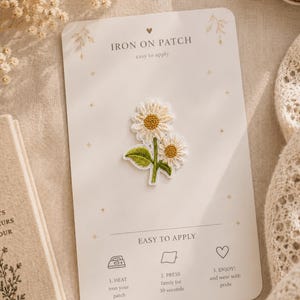 Peut inclure: Un écusson thermocollant brodé représentant deux marguerites blanches aux cœurs jaunes avec des tiges et des feuilles vertes. L'écusson est fixé sur une carte rectangulaire blanche portant le texte IRON ON PATCH et des instructions.