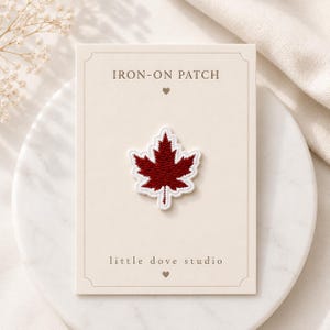 May include: An iron-on patch shaped like a maple leaf with deep red embroidery and a white border. The patch is mounted on a rectangular cream-coloured card labelled IRON-ON PATCH and little dove studio with small heart icons above and below.