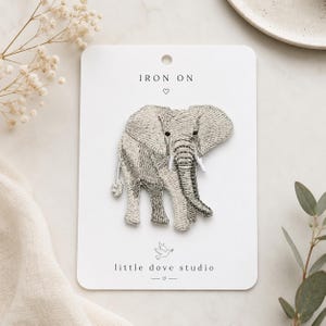 May include: An iron-on embroidered patch featuring a detailed elephant in shades of grey and white. The patch is mounted on a white rectangular display card with the text IRON ON and little dove studio printed in black ink with small heart icons.
