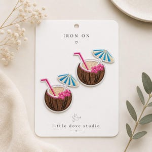 May include: Two embroidered iron-on patches featuring brown coconuts with tan centers, pink straws, blue and white striped umbrellas, and small pink flowers. The patches are displayed on a white card labeled IRON ON and little dove studio.