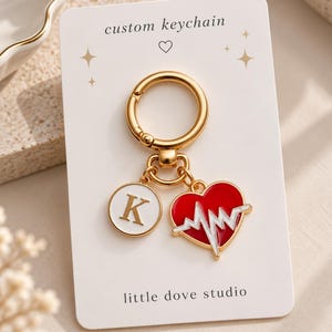 May include: A gold-toned metal keychain on a white display card labelled custom keychain and little dove studio. The keychain features a circular charm with the letter K in white and gold, and a red heart charm with a white EKG heartbeat line.