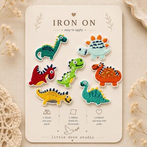 May include: A set of seven embroidered dinosaur patches on a beige card labelled IRON ON. The patches feature various dinosaurs in green, red, orange, and yellow. The card includes instructions: 1. HEAT iron your patch, 2. PRESS firmly for 30 seconds, 3. ENJOY! and wear with pride. The brand name little dove studio is printed at the bottom.