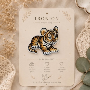 Peut inclure: Un écusson thermocollant brodé représentant un petit tigre avec un pelage orange, noir et blanc et des yeux bleus. L'écusson est fixé sur une carte beige avec le texte IRON ON, des instructions d'application et la marque little dove studio.