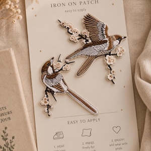 Puede incluir: Dos parches bordados termoadhesivos con pájaros marrones y blancos posados en ramas florecientes. Los parches se presentan en una tarjeta de color crema con el texto "IRON ON PATCH" y "EASY TO APPLY" y tres pasos numerados de aplicación.