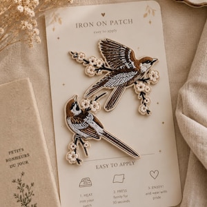 May include: Two embroidered iron-on patches featuring brown and white birds perched on flowering branches. The patches are displayed on a cream-colored backing card with instructions labeled 1. HEAT, 2. PRESS, and 3. ENJOY! and wear with pride.
