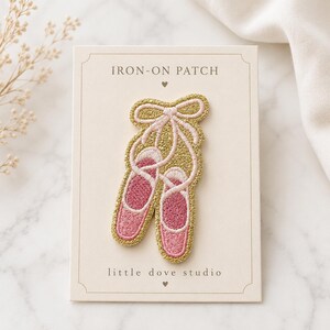 May include: An iron-on patch featuring a pair of pink ballet slippers with gold glitter embroidery on a rectangular card labeled IRON-ON PATCH and little dove studio. The slippers are tied together with a gold glitter bow at the top.