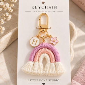 May include: A macrame rainbow keyring featuring purple, pink, and cream yarn arches with cream tassels. It includes a gold-tone lobster clasp, a white circular charm with the letter H, and a small pink flower charm on a white display card.