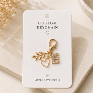 May include: A gold-toned keychain featuring a lobster clasp, a leaf charm, a beaded heart charm, and a letter E charm encrusted with clear crystals. The keychain is attached to a white display card printed with the text CUSTOM KEYCHAIN and LITTLE DOVE STUDIO.