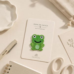 May include: An embroidered iron-on patch shaped like a green frog with a light green belly, black eyes, and a smiling mouth. The patch is attached to a rectangular white card featuring the text IRON ON PATCH, EASY TO IRON ON, and MADE WITH LOVE.