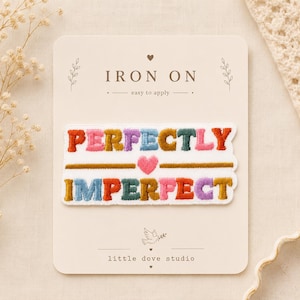 Peut inclure: Un écusson brodé thermocollant sur une carte de présentation. L'écusson affiche le texte 'PERFECTLY IMPERFECT' en lettres brodées multicolores en relief avec un petit cœur rose au centre. La carte porte les mentions 'IRON ON' et 'little dove studio'.