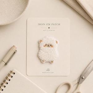 May include: A white, fluffy, embroidered iron-on patch shaped like a small sheep on a rectangular display card. The card features the text IRON ON PATCH, EASY TO IRON ON, and MADE WITH LOVE, along with a small iron icon and a heart symbol.
