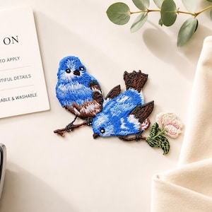 May include: An embroidered patch featuring two blue birds with brown wings perched on a branch with a small pink flower. Next to the patch is an instruction card labelled IRON ON, EASY TO APPLY, BEAUTIFUL DETAILS, and DURABLE & WASHABLE.