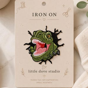Peut inclure: Un écusson thermocollant brodé représentant une tête de dinosaure verte avec la gueule ouverte et des dents pointues, émergeant d'un trou noir dentelé. L'écusson est fixé sur une carte de présentation beige marquée IRON ON et little dove studio.