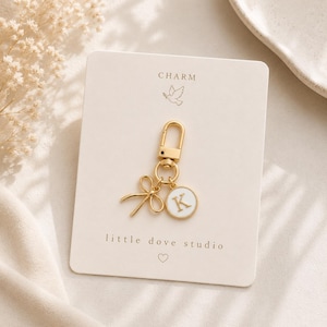 May include: A gold-toned metal charm featuring a lobster clasp, a small bow, and a circular pendant with a white enamel inlay and the letter K in gold. The charm is attached to a cream-coloured display card printed with the text CHARM, little dove studio, and a small heart icon.