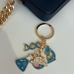 Puede incluir: Un llavero dorado con múltiples colgantes con temática científica. Los colgantes incluyen una cadena de ADN, un matraz, un microscopio y un globo terráqueo con el texto "CHEMISTRY". Los colgantes son azules, rosas y dorados. También se ven una cadena dorada y un bolso azul marino.