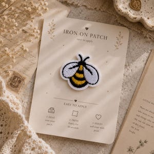 May include: An embroidered bee iron-on patch with yellow and black stripes and white wings, attached to a cream-coloured backing card. The card features the text IRON ON PATCH, EASY TO APPLY, and instructions for heating, pressing for 30 seconds, and wearing.
