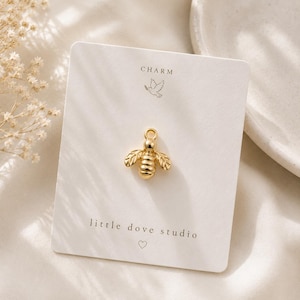 May include: A gold-toned bee charm centered on a rectangular white display card. The card features the text CHARM at the top, a small bird icon, and the text little dove studio with a heart symbol at the bottom. The bee has detailed wings.