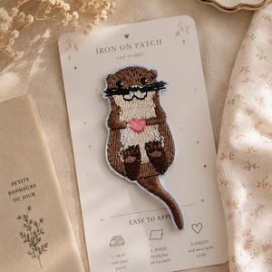May include: An embroidered iron-on patch featuring a brown and cream-coloured otter holding a small pink heart. The patch is attached to a white rectangular backing card with the text IRON ON PATCH, EASY TO APPLY, and instructions for use.