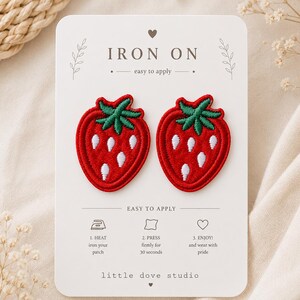 May include: Two red strawberry embroidered patches with green leaves and white seeds on a white display card. The card features the text IRON ON, instructions for application, and the brand name little dove studio at the bottom.