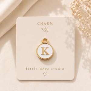 May include: A gold-toned circular charm featuring a white enamel inlay with the letter K in the center. The charm is attached to a beige display card printed with the text CHARM, a dove icon, little dove studio, and a small heart icon.