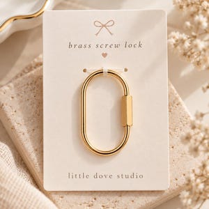 Peut inclure: Un mousqueton ovale en laiton doré avec un mécanisme de verrouillage à vis, fixé sur une carte de présentation rectangulaire. La carte porte une icône de nœud, le texte "brass screw lock" avec un petit cœur et "little dove studio".