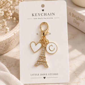Peut inclure: Un porte-clés doré sur une carte de présentation blanche avec le texte KEYCHAIN little details, big meaning. et LITTLE DOVE STUDIO. Le porte-clés comprend une breloque tour Eiffel dorée avec des cristaux, un cœur blanc et un disque blanc avec la lettre C.