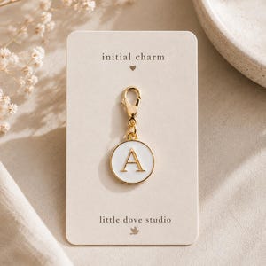 Puede incluir: Un dije circular dorado con incrustación de esmalte blanco y la letra A en oro. El dije incluye un cierre de mosquetón y está sujeto a una tarjeta de presentación rectangular impresa con el texto initial charm y little dove studio.